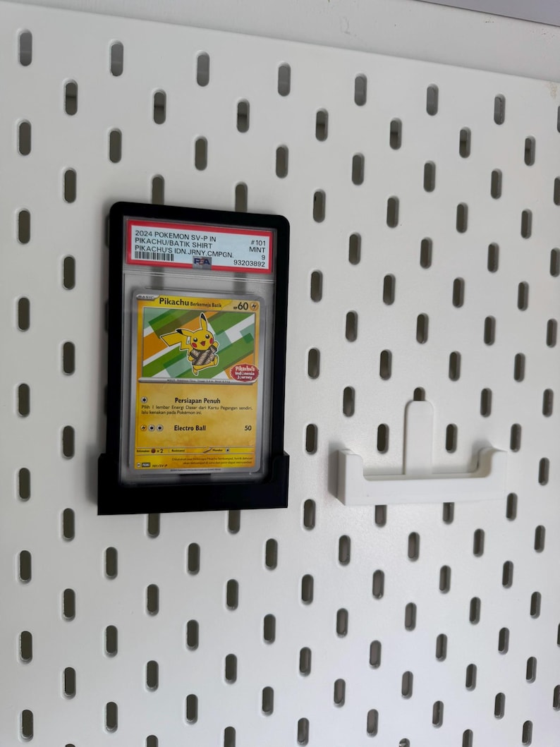 May include: A framed Pok&eacute;mon trading card featuring Pikachu, displayed on a white pegboard. The card is in a black holder and includes text such as "2024 Pokemon SV-P" and "Pikachu Batik Shirt". A white pegboard hook is also visible.