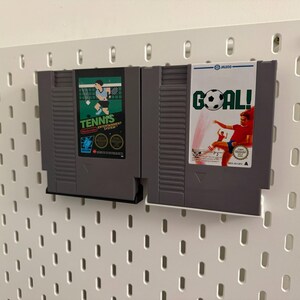 Pode incluir: Dois cartuchos de jogos cinzentos do Nintendo Entertainment System. Um apresenta um gr&aacute;fico de jogador de t&eacute;nis com a palavra "TENNIS". O outro tem um gr&aacute;fico de jogador de futebol com a palavra "GOAL!". Ambos est&atilde;o montados numa placa perfurada branca.