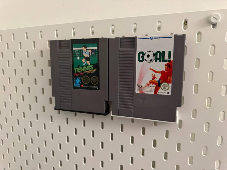 Pode incluir: Dois cartuchos de jogos cinzentos do Nintendo Entertainment System. Um apresenta um gr&aacute;fico de jogador de t&eacute;nis com a palavra "TENNIS". O outro tem um gr&aacute;fico de jogador de futebol com a palavra "GOAL!". Ambos est&atilde;o montados numa placa perfurada branca.