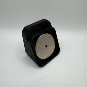 May include: A black, 3D-printed magnetic phone mount. The mount has a square base with a curved front and a circular silver magnet. The magnet has a small hole in the centre. The mount is designed to hold a phone.