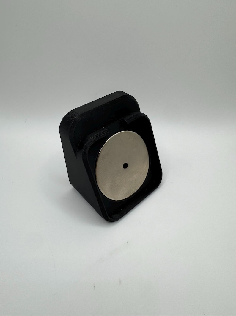 May include: A black, 3D-printed magnetic phone mount. The mount has a square base with a curved front and a circular silver magnet. The magnet has a small hole in the centre. The mount is designed to hold a phone.
