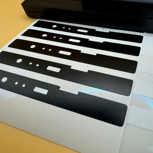 May include: A sheet of white paper with six black vinyl stickers. The stickers are rectangular and have a textured surface. The stickers are arranged in a row, with the largest sticker at the bottom and the smallest sticker at the top.