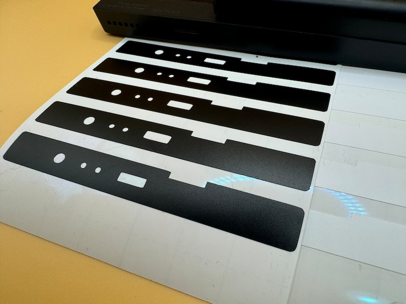 May include: A sheet of white paper with six black vinyl stickers. The stickers are rectangular and have a textured surface. The stickers are arranged in a row, with the largest sticker at the bottom and the smallest sticker at the top.