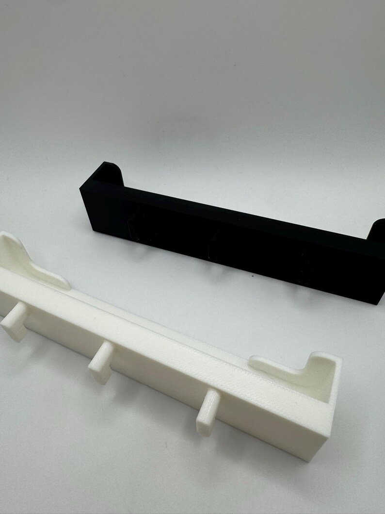 May include: Two rectangular shelves, one black and one white, are displayed against a white background. The white shelf has several small, vertical protrusions. The black shelf is a solid, simple design. Both shelves have a minimalist aesthetic.