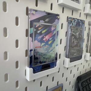 K&ouml;nnte beinhalten: Eine wei&szlig;e Lochwand mit mehreren durchsichtigen Plastikkartenhaltern, die farbige Sammelkarten zeigen. Die Karten zeigen Illustrationen von Charakteren und Szenen aus einem beliebten Sammelkartenspiel. Die Lochwand ist eine gro&szlig;artige M&ouml;glichkeit, Sammelkarten zu pr&auml;sentieren und zu organisieren.