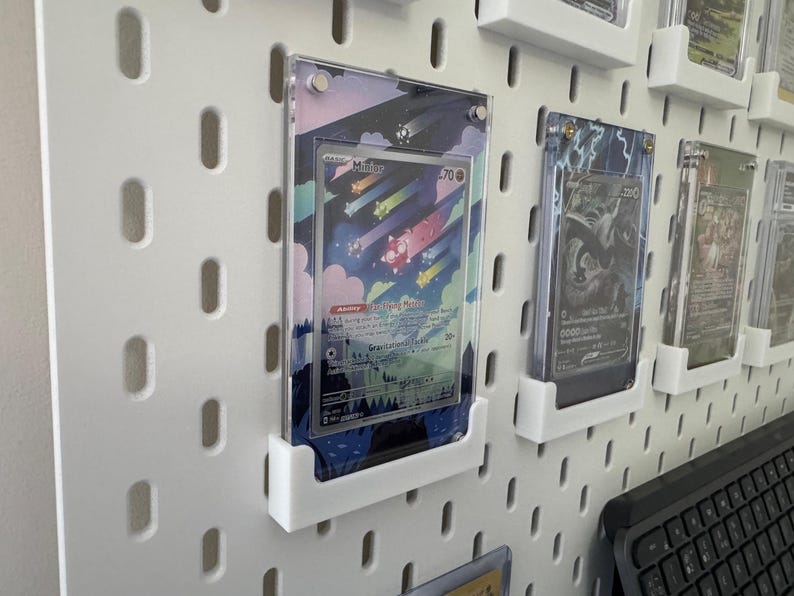 K&ouml;nnte beinhalten: Eine wei&szlig;e Lochwand mit mehreren durchsichtigen Plastikkartenhaltern, die farbige Sammelkarten zeigen. Die Karten zeigen Illustrationen von Charakteren und Szenen aus einem beliebten Sammelkartenspiel. Die Lochwand ist eine gro&szlig;artige M&ouml;glichkeit, Sammelkarten zu pr&auml;sentieren und zu organisieren.