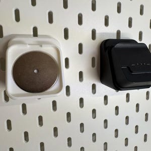 May include: Two wall-mounted storage containers. One is white with a circular, metallic object inside. The other is black and has a rectangular shape with a small lip. Both are mounted on a white pegboard.