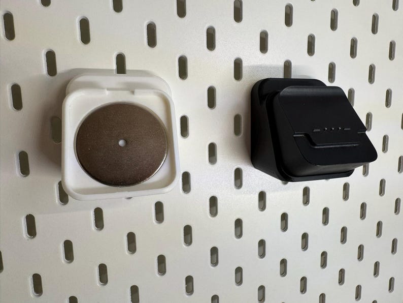 May include: Two wall-mounted storage containers. One is white with a circular, metallic object inside. The other is black and has a rectangular shape with a small lip. Both are mounted on a white pegboard.