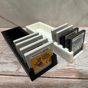 Puede incluir: Soportes de cartuchos de juego impresos en 3D en blanco y negro. Se muestran un soporte negro y un soporte blanco, cada uno con varios cartuchos de juego de Nintendo 3DS y DS. El juego Super Mario Maker 3DS es visible.