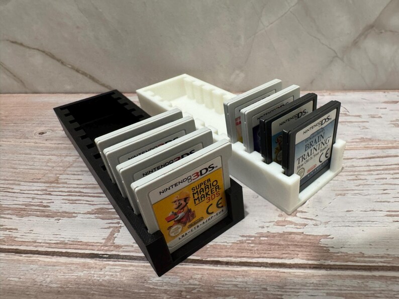Puede incluir: Soportes de cartuchos de juego impresos en 3D en blanco y negro. Se muestran un soporte negro y un soporte blanco, cada uno con varios cartuchos de juego de Nintendo 3DS y DS. El juego Super Mario Maker 3DS es visible.