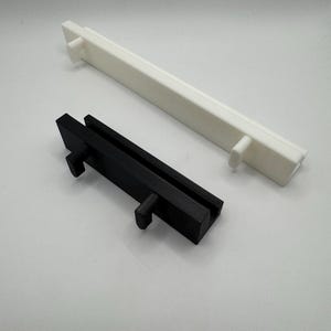 以下が含まれることがあります： 2つの長方形の3Dプリントオブジェクト。1つは黒、もう1つは白です。どちらも長方形のベースと両端に突き出たペグを備えた同様のデザインです。黒いオブジェクトは白いオブジェクトよりも短いです。