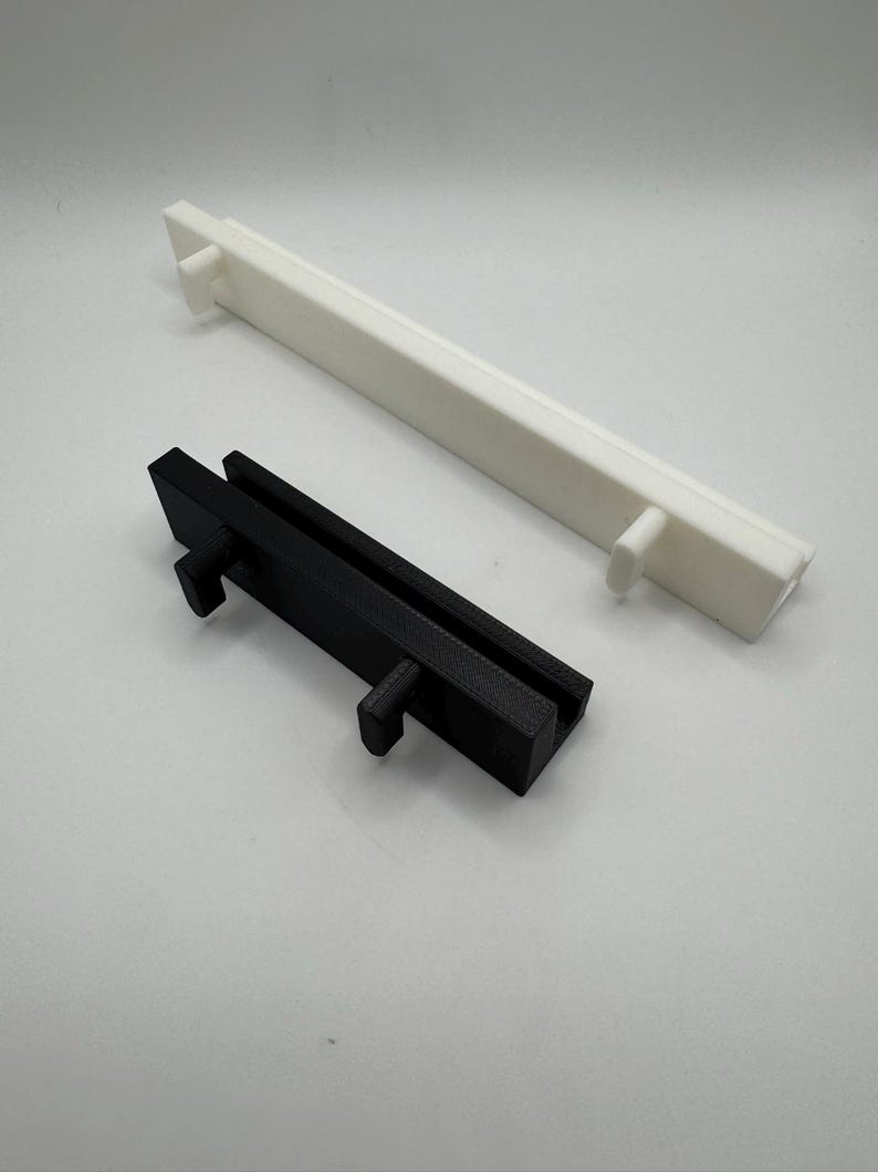 以下が含まれることがあります： 2つの長方形の3Dプリントオブジェクト。1つは黒、もう1つは白です。どちらも長方形のベースと両端に突き出たペグを備えた同様のデザインです。黒いオブジェクトは白いオブジェクトよりも短いです。