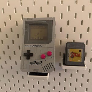 May include: A gray Nintendo Game Boy with pink buttons and a game cartridge, mounted on a white pegboard. The Game Boy screen displays a pixelated game scene. A Zelda game cartridge is also displayed.