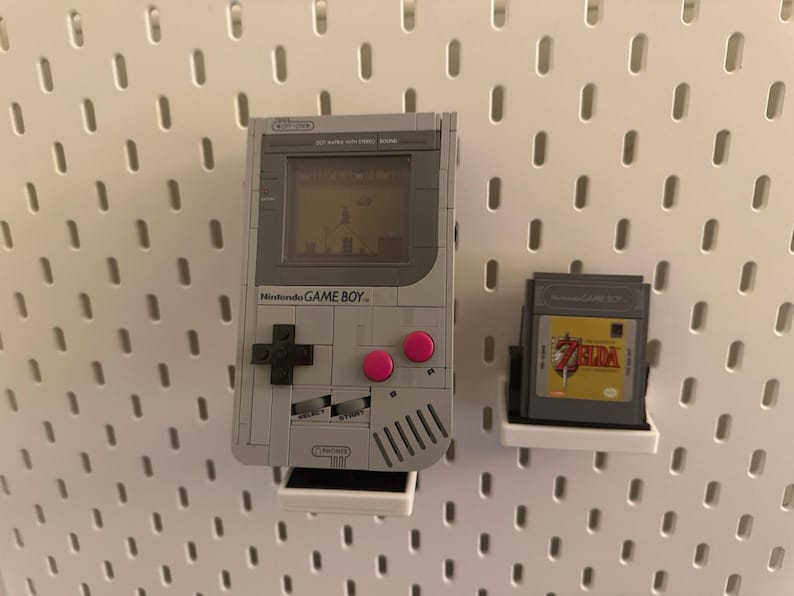 May include: A gray Nintendo Game Boy with pink buttons and a game cartridge, mounted on a white pegboard. The Game Boy screen displays a pixelated game scene. A Zelda game cartridge is also displayed.