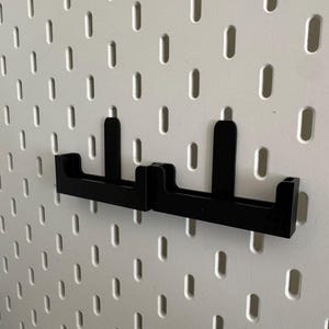 May include: Two black pegboard hooks mounted on a white pegboard. The hooks are made of a solid material and have a simple, functional design. The pegboard has evenly spaced oval holes.