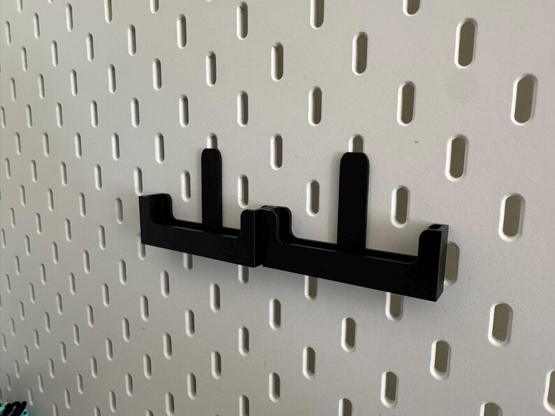 May include: Two black pegboard hooks mounted on a white pegboard. The hooks are made of a solid material and have a simple, functional design. The pegboard has evenly spaced oval holes.