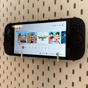 May include: A black handheld gaming console mounted on a white pegboard. The screen displays a game menu with several game icons. The console is held in place by white brackets.