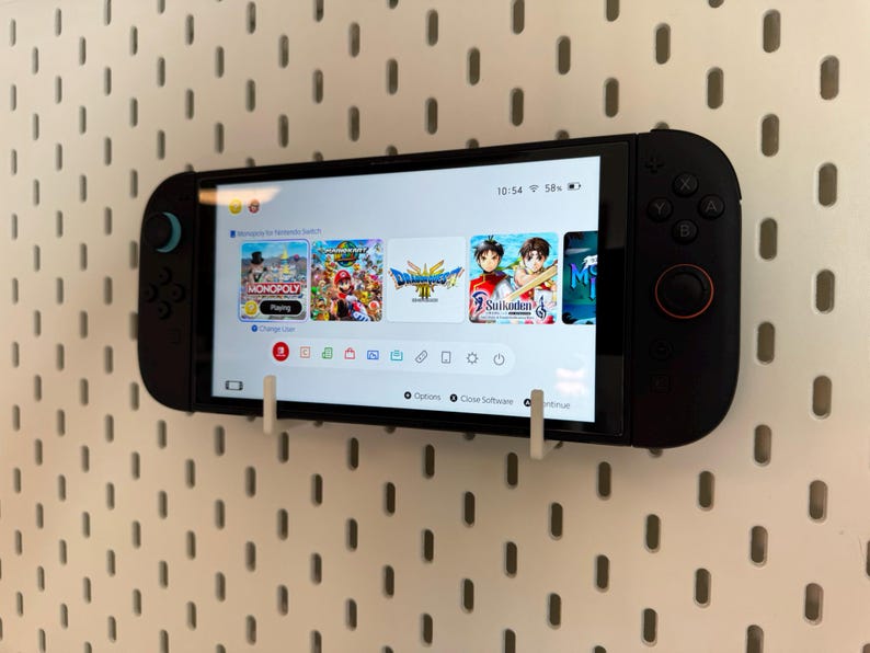 May include: A black handheld gaming console mounted on a white pegboard. The screen displays a game menu with several game icons. The console is held in place by white brackets.
