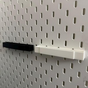 K&ouml;nnte beinhalten: Zwei rechteckige Regale, eines schwarz und eines wei&szlig;, an einer wei&szlig;en Lochwand befestigt. Die Regale sind aus Kunststoff und f&uuml;r kleine Gegenst&auml;nde konzipiert. Die Lochwand hat ein Gitter aus gleichm&auml;&szlig;ig verteilten L&ouml;chern.
