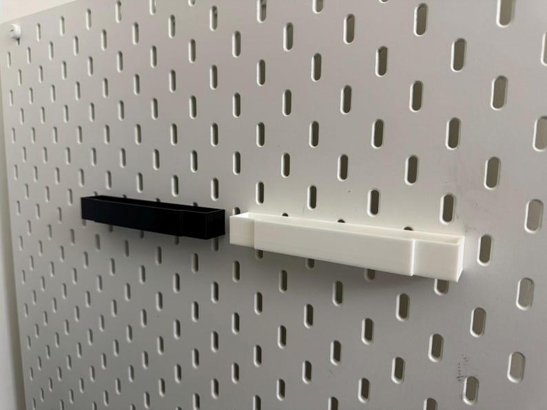 K&ouml;nnte beinhalten: Zwei rechteckige Regale, eines schwarz und eines wei&szlig;, an einer wei&szlig;en Lochwand befestigt. Die Regale sind aus Kunststoff und f&uuml;r kleine Gegenst&auml;nde konzipiert. Die Lochwand hat ein Gitter aus gleichm&auml;&szlig;ig verteilten L&ouml;chern.