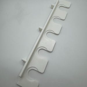 May include: A white, 3D-printed wall-mounted rod holder with five curved slots. The holder has a rectangular back with small pegs for mounting. The slots are designed to hold rods or similar items. The holder is made of plastic.