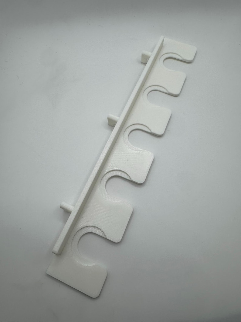 May include: A white, 3D-printed wall-mounted rod holder with five curved slots. The holder has a rectangular back with small pegs for mounting. The slots are designed to hold rods or similar items. The holder is made of plastic.