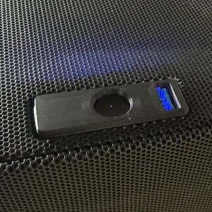 Op de afbeelding: Een zwarte luidspreker met een zwart metalen rooster en een kleine zwarte rechthoekige knop met een blauw licht.