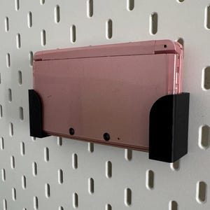 Pode incluir: Um console de jogos port&aacute;til Nintendo 3DS rosa est&aacute; montado em um suporte de parede preto. O suporte est&aacute; fixado em uma parede de placa de pinos branca.