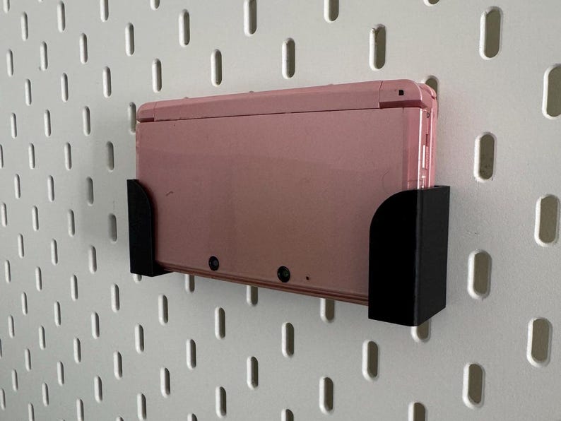 Pode incluir: Um console de jogos port&aacute;til Nintendo 3DS rosa est&aacute; montado em um suporte de parede preto. O suporte est&aacute; fixado em uma parede de placa de pinos branca.