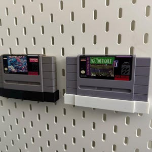 K&ouml;nnte beinhalten: Zwei graue Super Nintendo-Spielmodule. Eines zeigt eine farbenfrohe Grafik und das Capcom-Logo. Das andere zeigt "PGA TOUR GOLF" mit einer Golfplatzszene. Beide sind an einer wei&szlig;en Lochwand befestigt.
