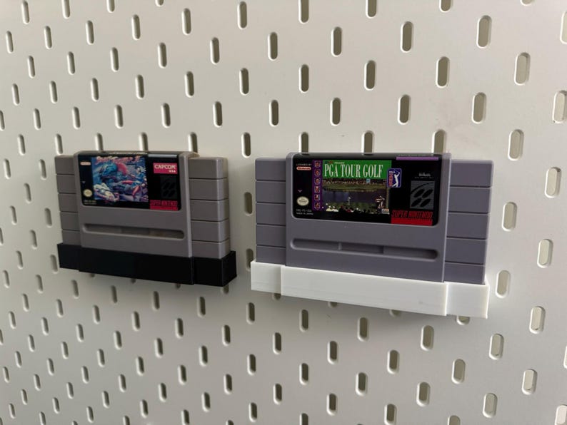 K&ouml;nnte beinhalten: Zwei graue Super Nintendo-Spielmodule. Eines zeigt eine farbenfrohe Grafik und das Capcom-Logo. Das andere zeigt "PGA TOUR GOLF" mit einer Golfplatzszene. Beide sind an einer wei&szlig;en Lochwand befestigt.