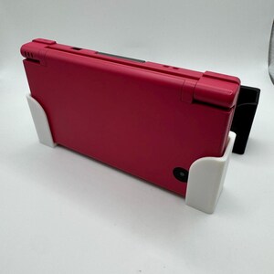 K&ouml;nnte beinhalten: Eine rote Handheld-Spielkonsole wird in einem wei&szlig;en und schwarzen St&auml;nder gehalten. Die Konsole ist geschlossen und zeigt ihre Oberseite. Der St&auml;nder ist so konzipiert, dass er die Konsole aufrecht h&auml;lt. Der Hintergrund ist schlicht wei&szlig;.