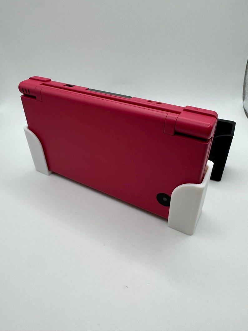 K&ouml;nnte beinhalten: Eine rote Handheld-Spielkonsole wird in einem wei&szlig;en und schwarzen St&auml;nder gehalten. Die Konsole ist geschlossen und zeigt ihre Oberseite. Der St&auml;nder ist so konzipiert, dass er die Konsole aufrecht h&auml;lt. Der Hintergrund ist schlicht wei&szlig;.