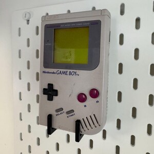 May include: A light gray Nintendo Game Boy handheld console with a black directional pad, purple buttons, and the text "Nintendo GAME BOY." The screen is yellow. The console is mounted on a white pegboard with black brackets.