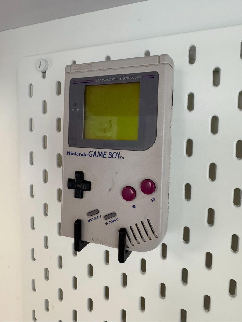 May include: A light gray Nintendo Game Boy handheld console with a black directional pad, purple buttons, and the text "Nintendo GAME BOY." The screen is yellow. The console is mounted on a white pegboard with black brackets.