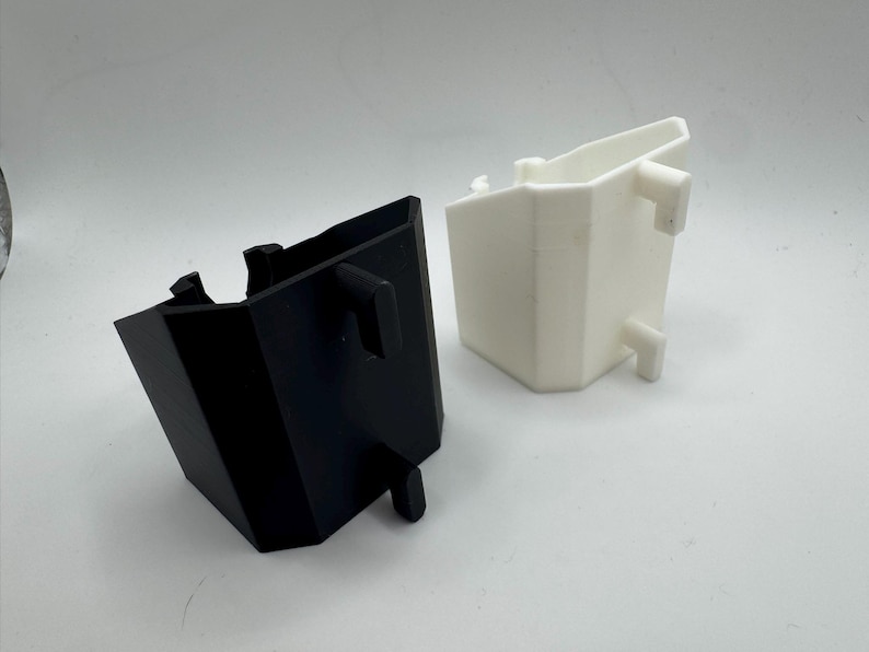 May include: Two plastic, geometric-shaped containers. One is black, the other is white. Both have a similar design with a rectangular body and protruding tabs. The containers are likely components of a larger product or system.