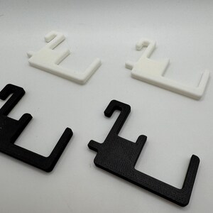 May include: Four 3D-printed plastic hangers. Two are white, and two are black. Each hanger has a hook and a rectangular base with a right angle. The hangers are designed to hold items.