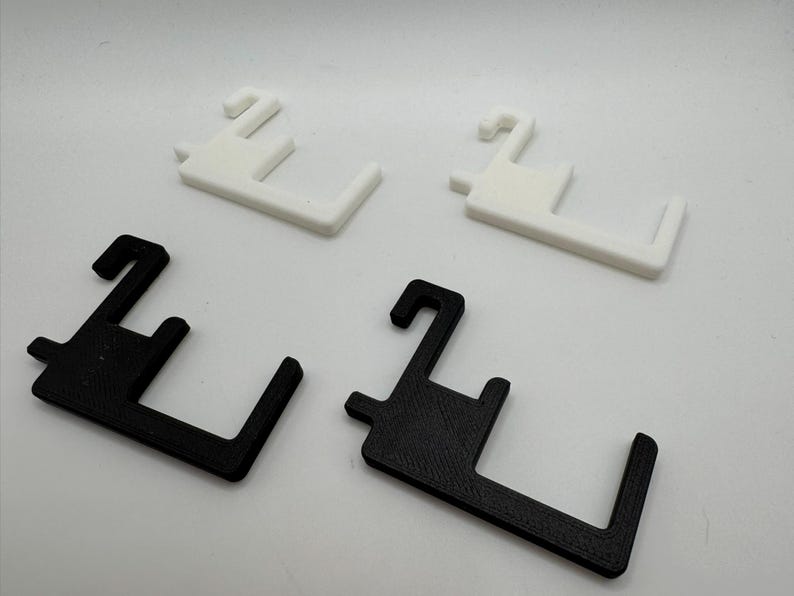 May include: Four 3D-printed plastic hangers. Two are white, and two are black. Each hanger has a hook and a rectangular base with a right angle. The hangers are designed to hold items.