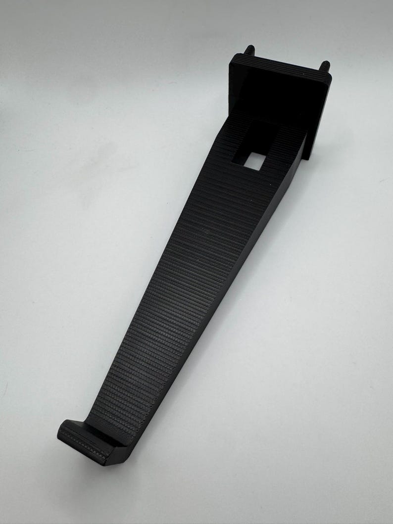 K&ouml;nnte beinhalten: Ein schwarzer, abgewinkelter Halter mit strukturierter Oberfl&auml;che. Der Halter hat eine quadratische Oberseite mit zwei kleinen Vorspr&uuml;ngen und einer rechteckigen &Ouml;ffnung. Die Unterseite hat einen kleinen, flachen Vorsprung. Der Halter dient zur Unterst&uuml;tzung eines Regals oder eines anderen Objekts.