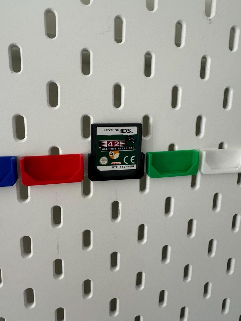 May include: A white pegboard with a Nintendo DS game cartridge in a green, red, and blue holder. The game cartridge is for 42 All-Time Classics.