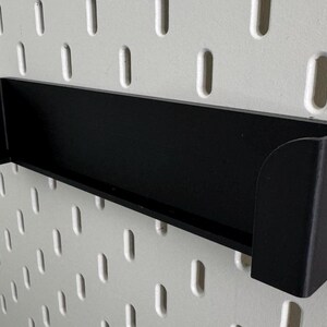 Op de afbeelding: Zwarte, wandgemonteerde plank van plastic met een afgeronde rand. De plank is gemonteerd op een witte pegboard-wand.