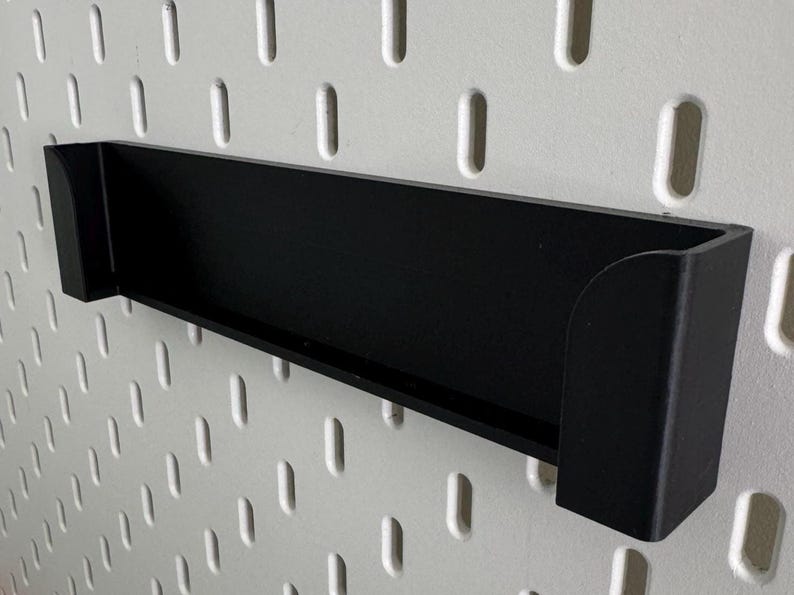 Op de afbeelding: Zwarte, wandgemonteerde plank van plastic met een afgeronde rand. De plank is gemonteerd op een witte pegboard-wand.