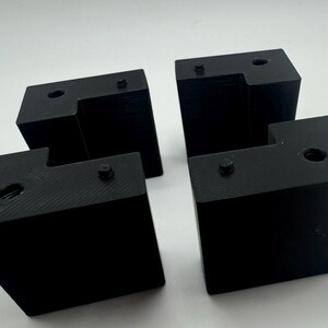 May include: Four black plastic corner brackets with holes for screws. The brackets are designed to connect two pieces of wood at a 90-degree angle.