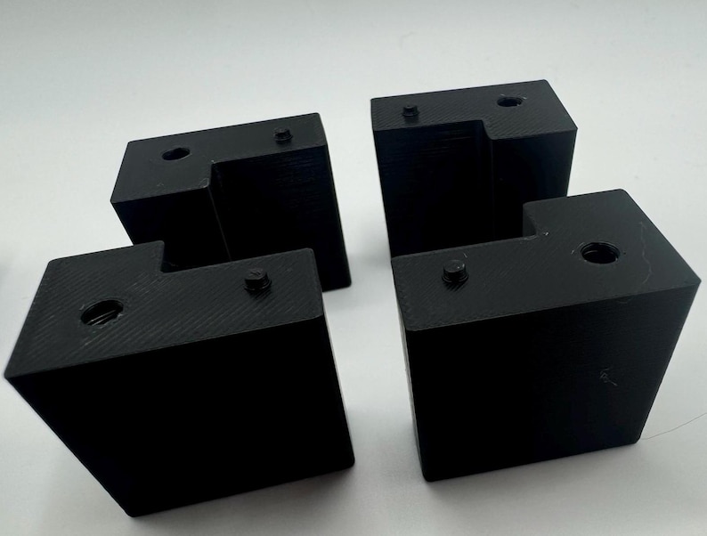 May include: Four black plastic corner brackets with holes for screws. The brackets are designed to connect two pieces of wood at a 90-degree angle.