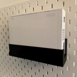 Peut inclure: Une console Nintendo Wii blanche mont&eacute;e sur une &eacute;tag&egrave;re noire, fix&eacute;e &agrave; une plaque perfor&eacute;e blanche. La console est centr&eacute;e, avec le logo Nintendo visible. La plaque perfor&eacute;e a des trous espac&eacute;s.