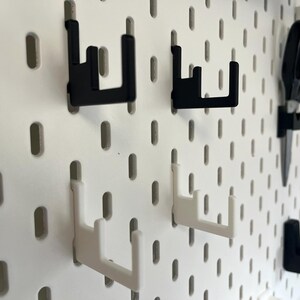 K&ouml;nnte beinhalten: Eine wei&szlig;e Lochwand mit schwarzen und wei&szlig;en Haken. Die Haken sind so konzipiert, dass sie in die L&ouml;cher der Lochwand passen und eine Aufbewahrungsl&ouml;sung bieten. Auch eine Schere ist zu sehen, was darauf hindeutet, dass die Lochwand zur Organisation von Werkzeugen verwendet wird.