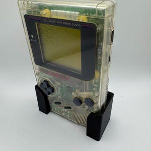 Op de afbeelding: Een doorzichtige, transparante Game Boy Color handheld-videogamesysteem met een zwart scherm en knoppen. De console staat in een zwarte standaard.