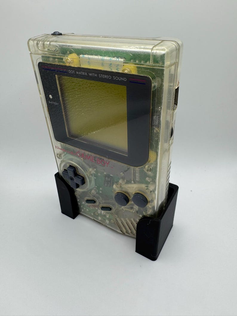 Op de afbeelding: Een doorzichtige, transparante Game Boy Color handheld-videogamesysteem met een zwart scherm en knoppen. De console staat in een zwarte standaard.