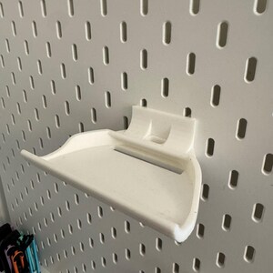 May include: A white, 3D-printed shelf designed to attach to a pegboard. The shelf has a curved front and a raised back with two small supports. The pegboard is white with evenly spaced oval holes.