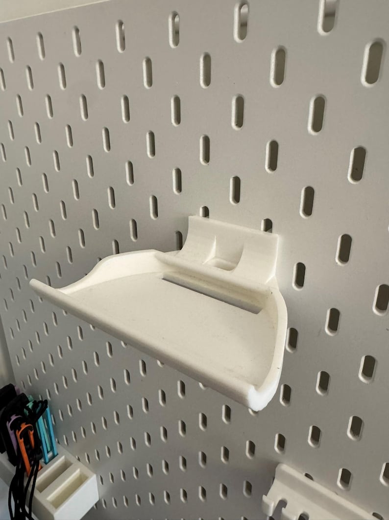 May include: A white, 3D-printed shelf designed to attach to a pegboard. The shelf has a curved front and a raised back with two small supports. The pegboard is white with evenly spaced oval holes.